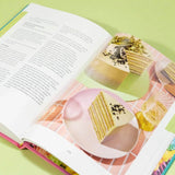 Open pages of The Crumbs & Doilies Recipe Book showing a layered cake recipe with full color photos.